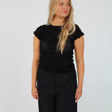En blond kvinde poserer i en sort, let gennemsigtig bluse og sorte bukser mod en hvid baggrund. Hun kigger til siden.