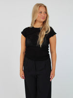 En blond kvinde poserer i en sort, let gennemsigtig bluse og sorte bukser mod en hvid baggrund. Hun kigger til siden.
