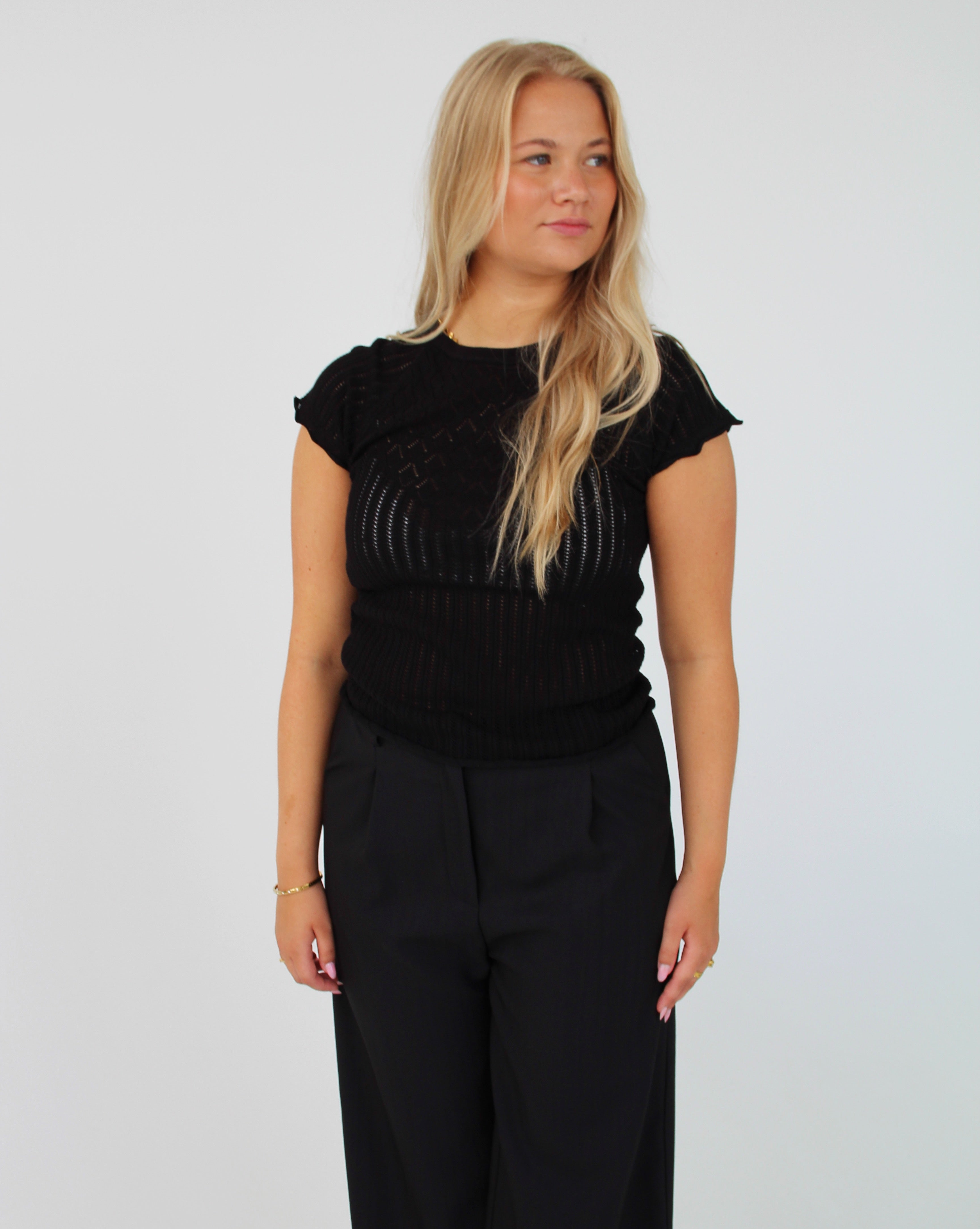 En blond kvinde poserer i en sort, let gennemsigtig bluse og sorte bukser mod en hvid baggrund. Hun kigger til siden.