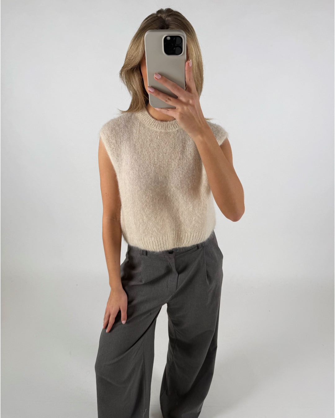 En kvinde tager en selfie med en telefon, der dækker hendes ansigt. Hun er iført en ærmeløs, creme-farvet striktrøje og grå bukser i et hvidt studie.
