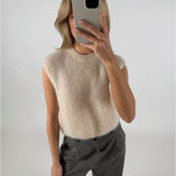 Person tager en selfie iført lyse ærmeløs sweater og grå bukser i et neutralt oplyst rum, dækkende ansigtet med en smartphone.