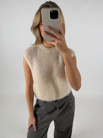 Person tager en selfie iført lyse ærmeløs sweater og grå bukser i et neutralt oplyst rum, dækkende ansigtet med en smartphone.