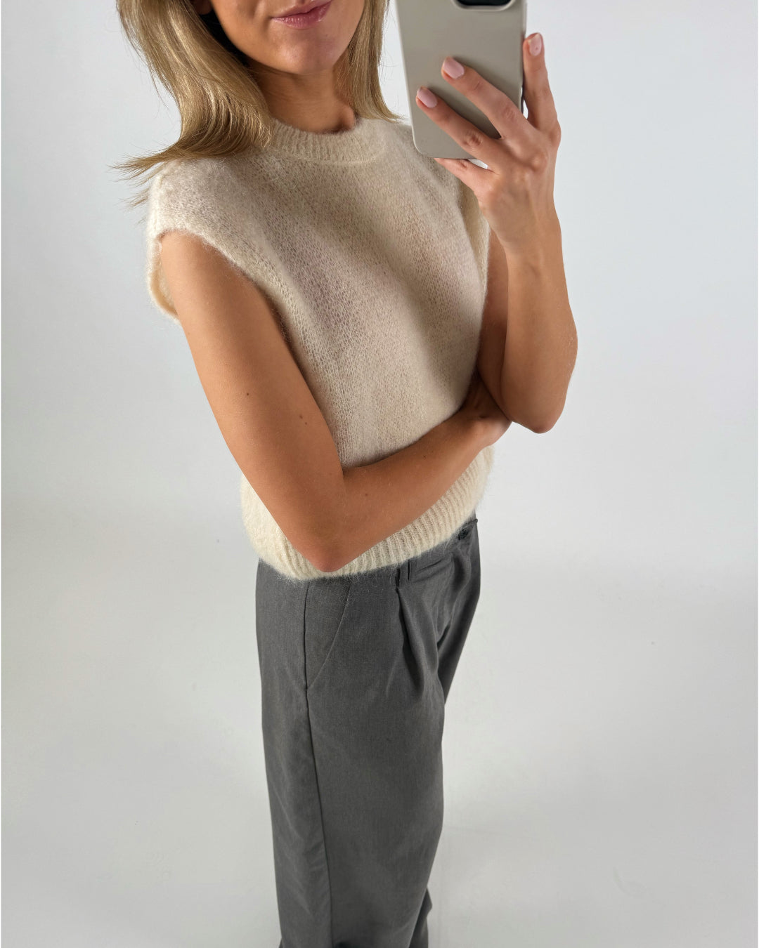 En kvinde tager en selfie med en smartphone. Hun er iført en ærmeløs, råhvid sweater og grå bukser foran en hvid baggrund.