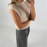En kvinde tager en selfie med en smartphone. Hun er iført en ærmeløs, råhvid sweater og grå bukser foran en hvid baggrund.