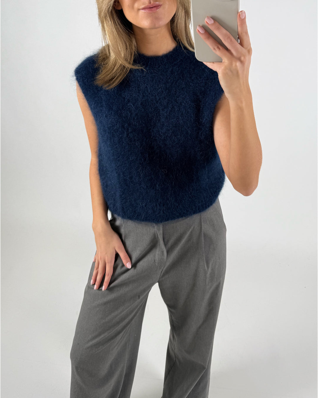 En kvinde tager en selfie, iført en mørkeblå, fuzzy vest og grå bukser. Hun står foran en hvid baggrund.