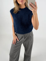 En kvinde tager en selfie, iført en mørkeblå, fuzzy vest og grå bukser. Hun står foran en hvid baggrund.