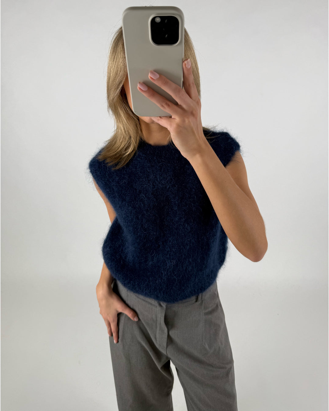 En kvinde tager et selfie med en lysegrå mobiltelefon foran hendes ansigt, iført en mørkeblå strikket vest og grå bukser i et hvidt studie.