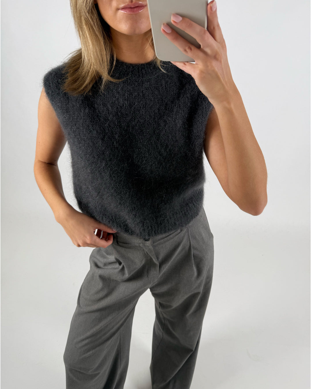 En kvinde tager et selfie med en telefon. Hun er iført en grå, fuzzy ærmeløs top og grå bukser mod en hvid baggrund.