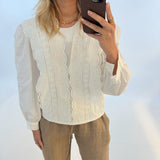 En kvinde tager en selfie med sin telefon. Hun er iført en hvid bluse med broderi og brune bukser, stående foran en hvid baggrund.