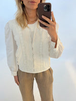 En kvinde tager en selfie med sin telefon. Hun er iført en hvid bluse med broderi og brune bukser, stående foran en hvid baggrund.