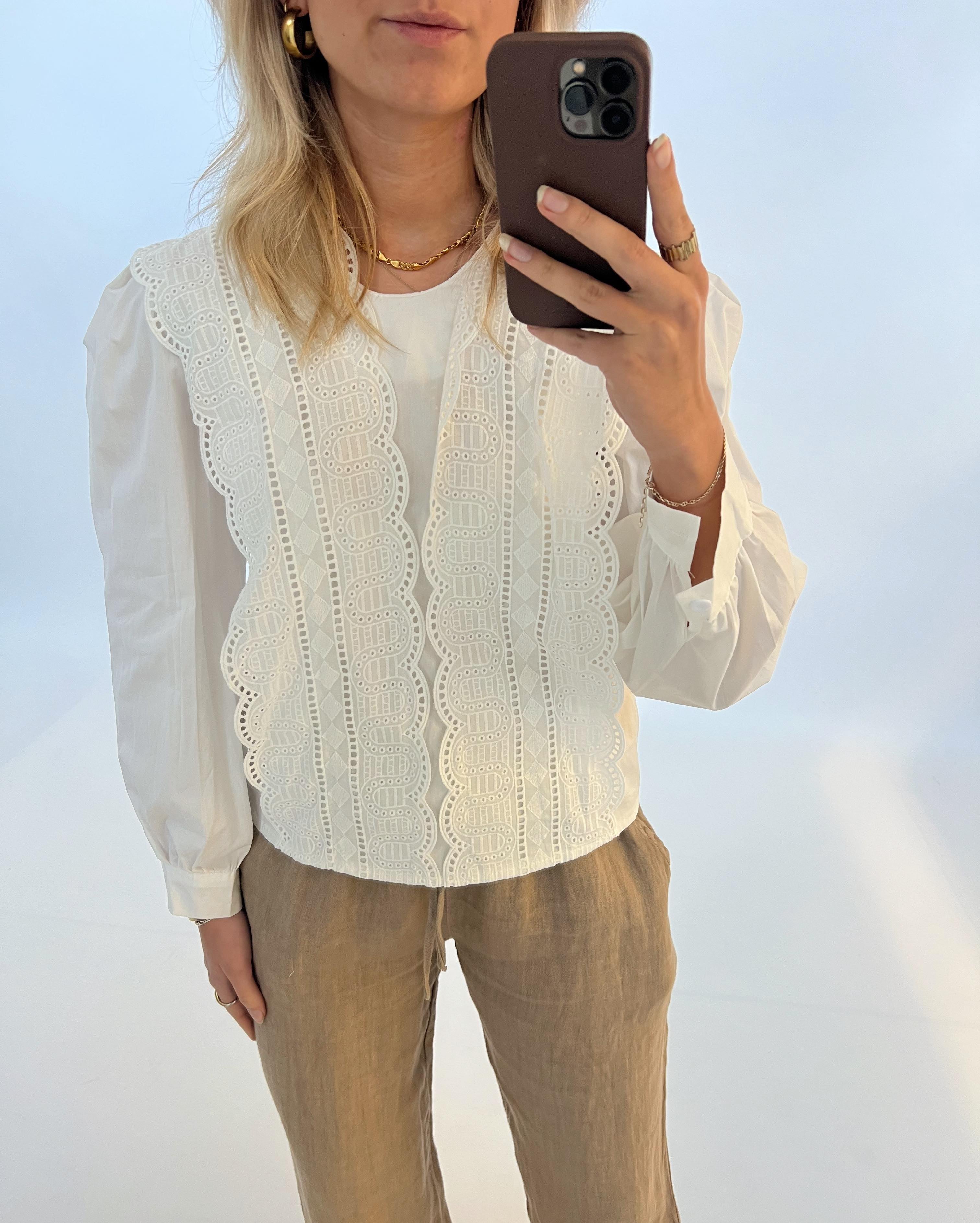 En kvinde tager en selfie med sin telefon. Hun er iført en hvid bluse med broderi og brune bukser, stående foran en hvid baggrund.