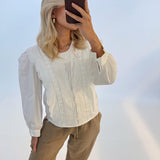 En kvinde holder en brun telefon op foran ansigtet i et lyst studie. Hun er iført en hvid bluse med blondedetaljer og brune bukser.