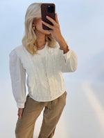 En kvinde holder en brun telefon op foran ansigtet i et lyst studie. Hun er iført en hvid bluse med blondedetaljer og brune bukser.