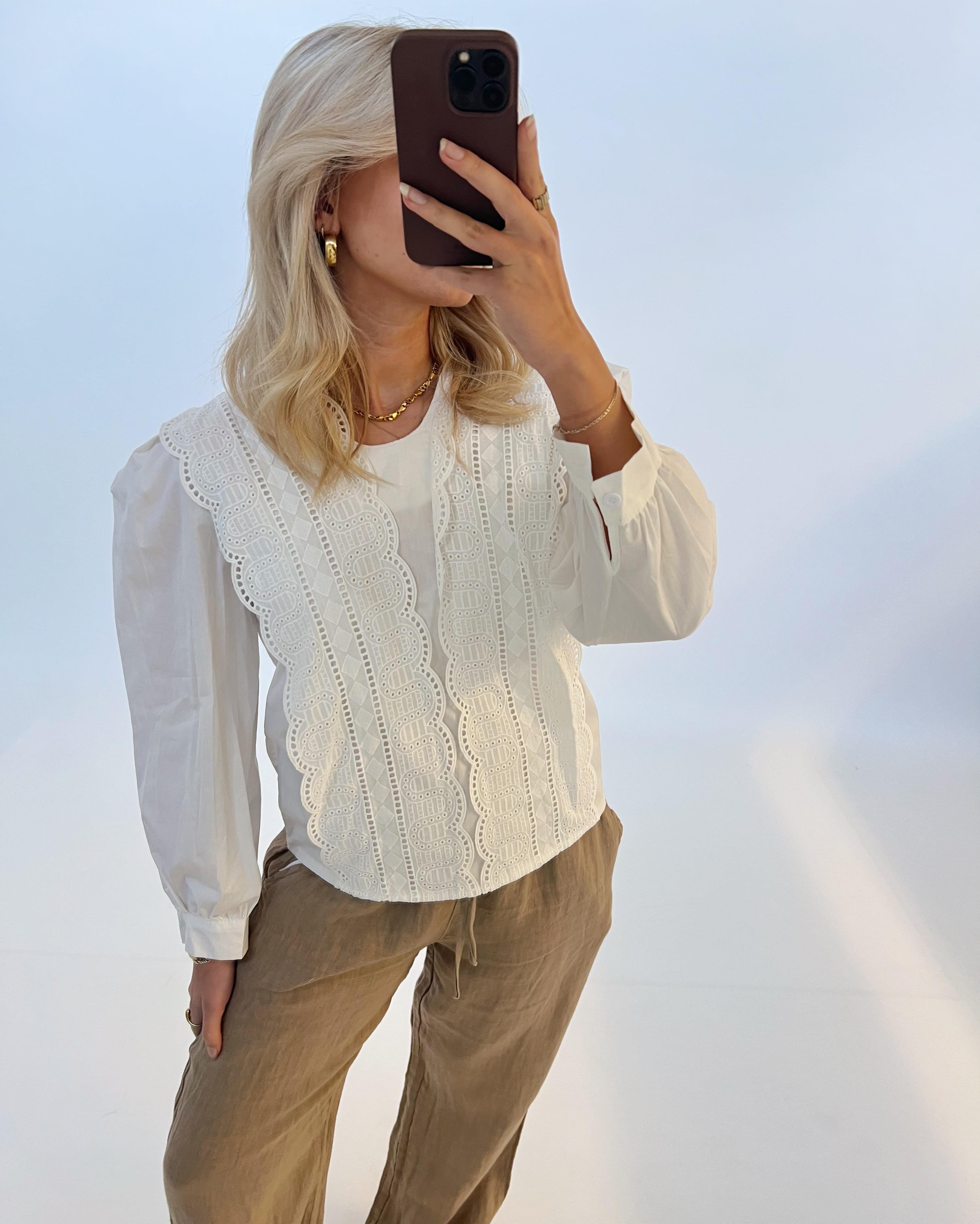 En kvinde holder en brun telefon op foran ansigtet i et lyst studie. Hun er iført en hvid bluse med blondedetaljer og brune bukser.