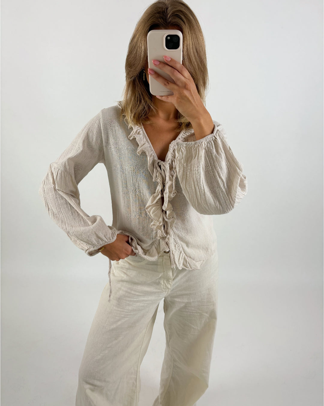 En kvinde tager et selfie med en telefon, hvor hendes ansigt er dækket af telefonen. Hun er iført en beige bluse med flæser og matchende bukser. Baggrunden er hvid.