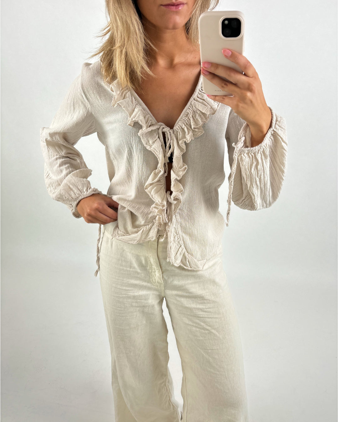 En kvinde tager et selfie med en telefon. Hun er iført en beige bluse med flæser og beige bukser, stående foran en hvid baggrund.