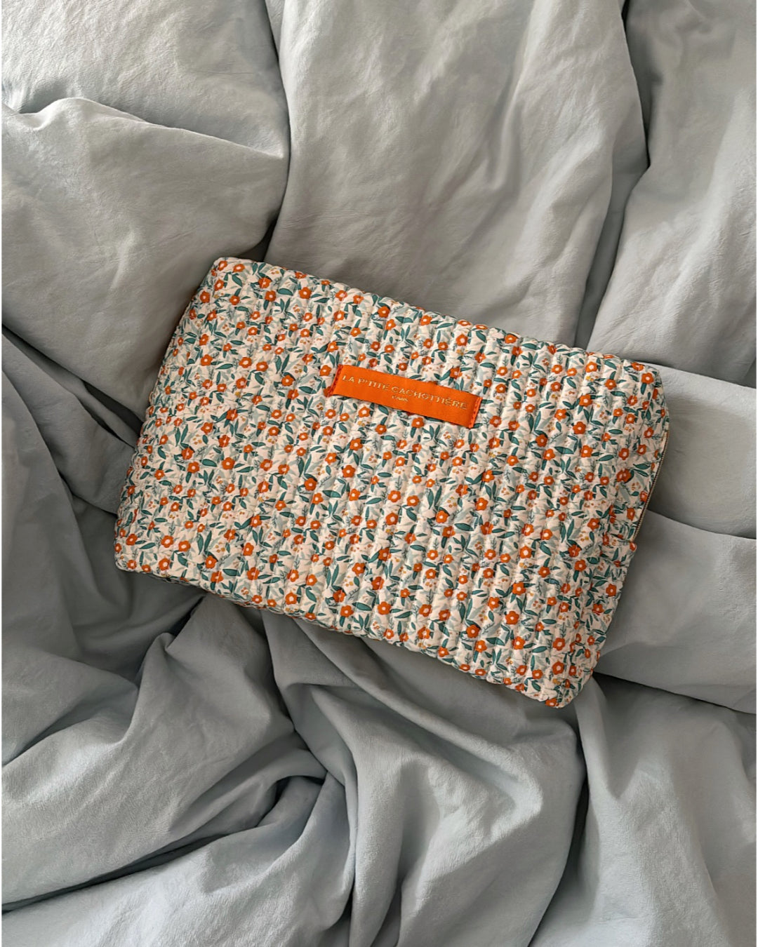 En blomstret, quiltet taske med et orange "LA PETITE CACHOTTIERE PARIS" mærke ligger på et lyseblåt, krøllet lagen.