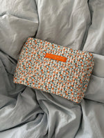 En blomstret, quiltet taske med et orange "LA PETITE CACHOTTIERE PARIS" mærke ligger på et lyseblåt, krøllet lagen.