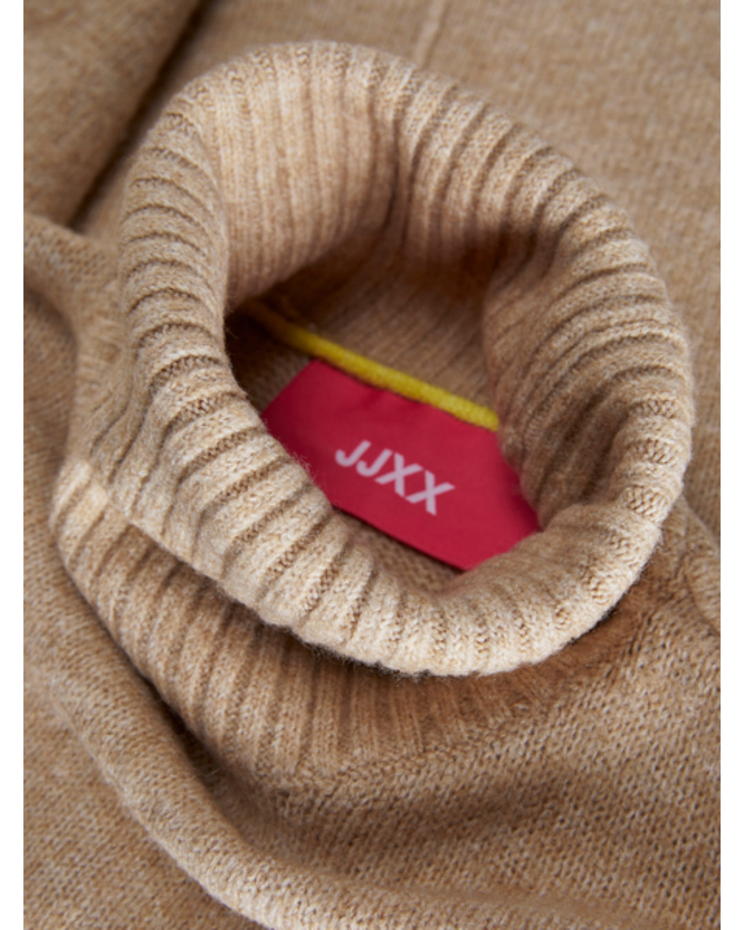 En lys brun rullekrave sweater folder sig, hvor en rød etiket med hvid tekst "JJXX" stikker ud fra kraven. En gul snor er også synlig.