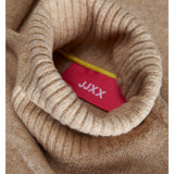 En lys brun rullekrave sweater folder sig, hvor en rød etiket med hvid tekst "JJXX" stikker ud fra kraven. En gul snor er også synlig.