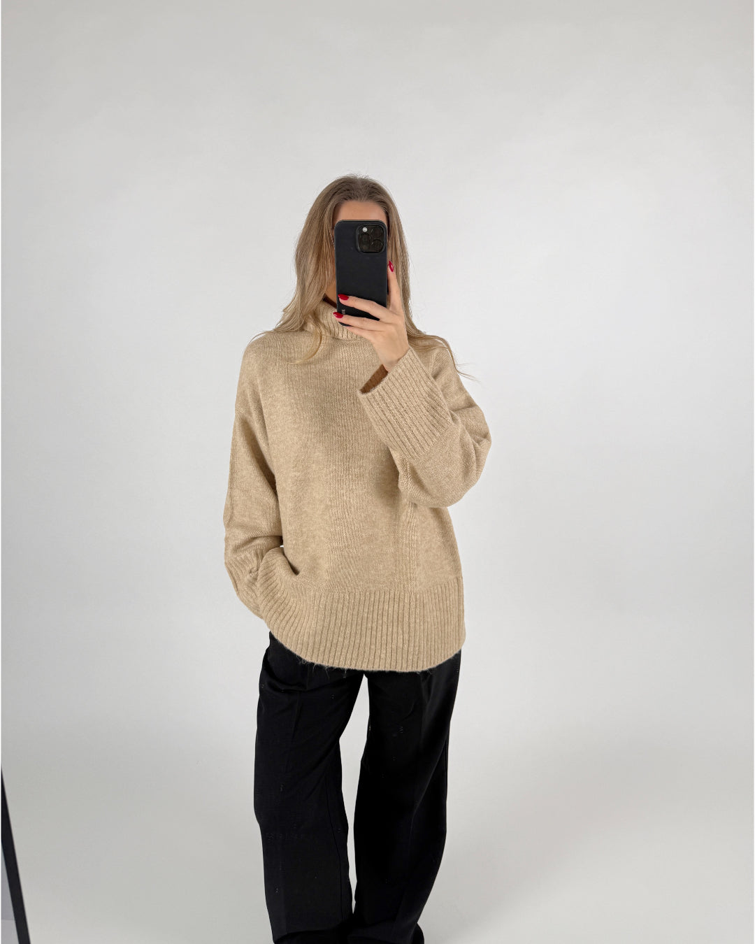 En person iført en beige sweater og sorte bukser tager et spejlbillede med en telefon i et neutralt, hvidt rum.