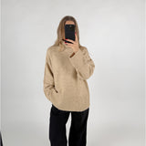 En person iført en beige sweater og sorte bukser tager et spejlbillede med en telefon i et neutralt, hvidt rum.