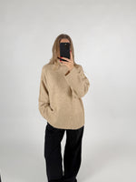 En person iført en beige sweater og sorte bukser tager et spejlbillede med en telefon i et neutralt, hvidt rum.