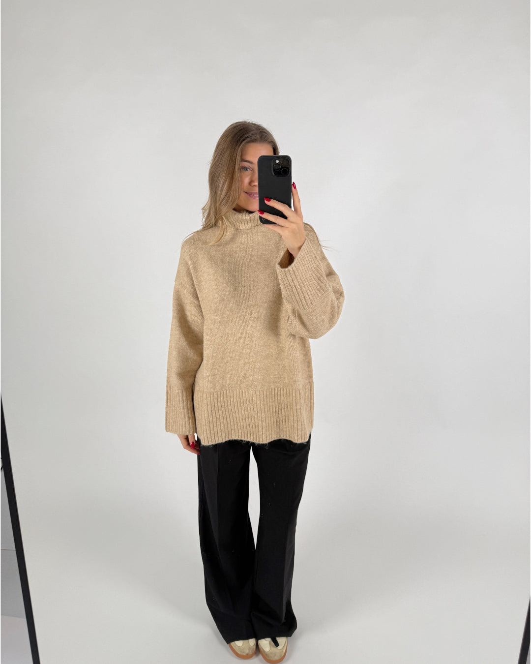 En kvinde tager et selfie med en telefon. Hun er iført en beige striktrøje og sorte bukser i et hvidt studie.