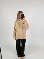 En kvinde tager et selfie med en telefon. Hun er iført en beige striktrøje og sorte bukser i et hvidt studie.