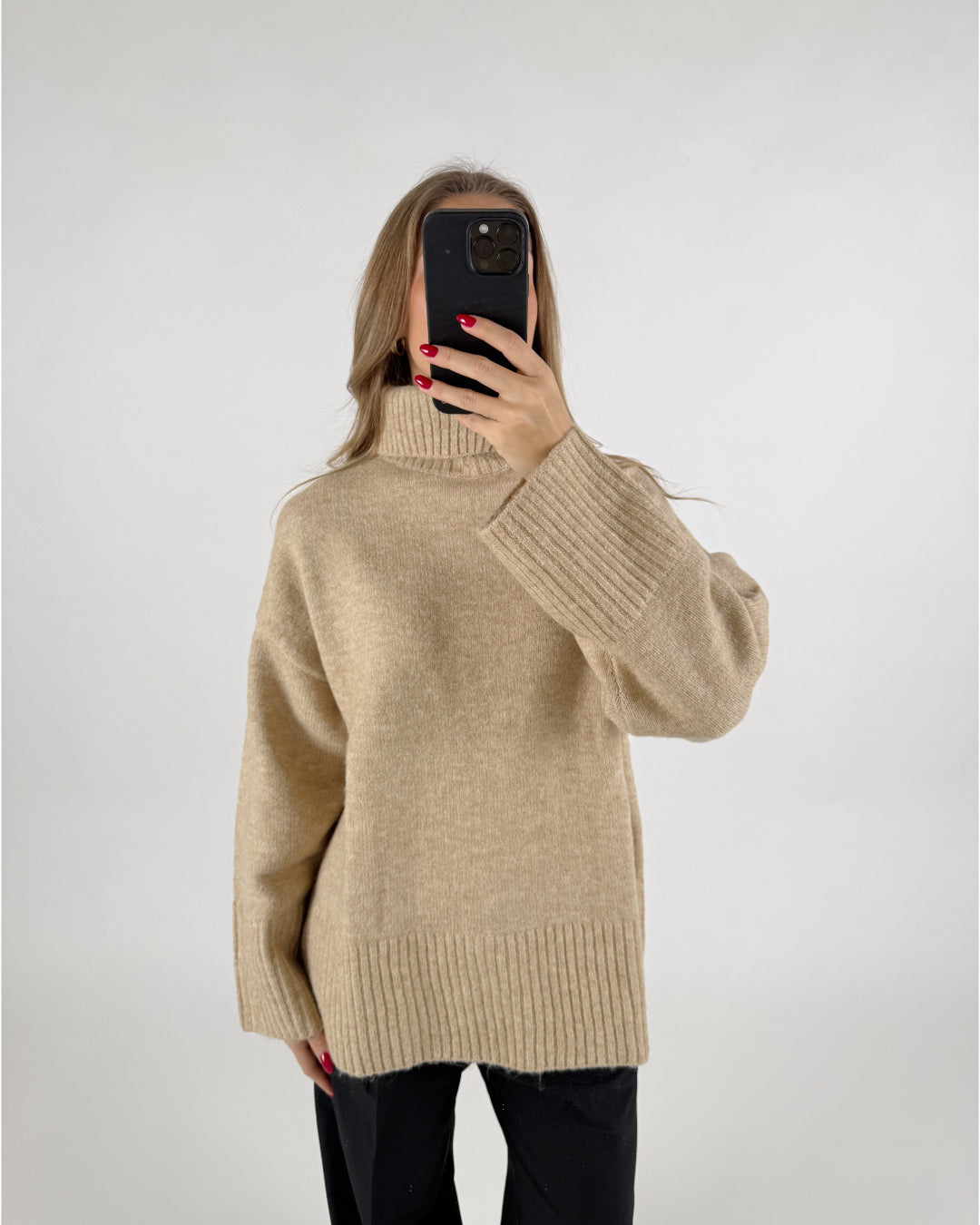 En kvinde tager et selfie med en sort mobiltelefon foran sit ansigt. Hun har en beige rullekrave sweater på og står foran en hvid baggrund.
