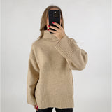 En kvinde tager et selfie med en sort mobiltelefon foran sit ansigt. Hun har en beige rullekrave sweater på og står foran en hvid baggrund.