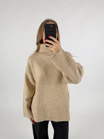 En kvinde tager et selfie med en sort mobiltelefon foran sit ansigt. Hun har en beige rullekrave sweater på og står foran en hvid baggrund.