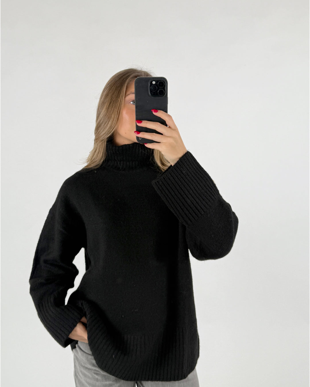 En kvinde holder en sort smartphone foran ansigtet i et selfie. Hun er iført en sort rullekrave og grå bukser, stående foran en hvid baggrund.