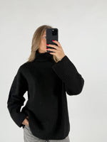En kvinde holder en sort smartphone foran ansigtet i et selfie. Hun er iført en sort rullekrave og grå bukser, stående foran en hvid baggrund.