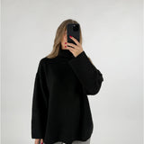 En kvinde holder en sort smartphone foran sit ansigt. Hun er iført en sort rullekrave sweater og grå bukser, i et hvidt studie.