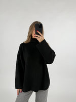 En kvinde holder en sort smartphone foran sit ansigt. Hun er iført en sort rullekrave sweater og grå bukser, i et hvidt studie.