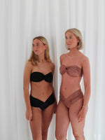 To blonde kvinder poserer i matchende bikinis foran et hvidt lagen. Den ene bærer en sort bikini, mens den anden bærer en mokkafarvet.