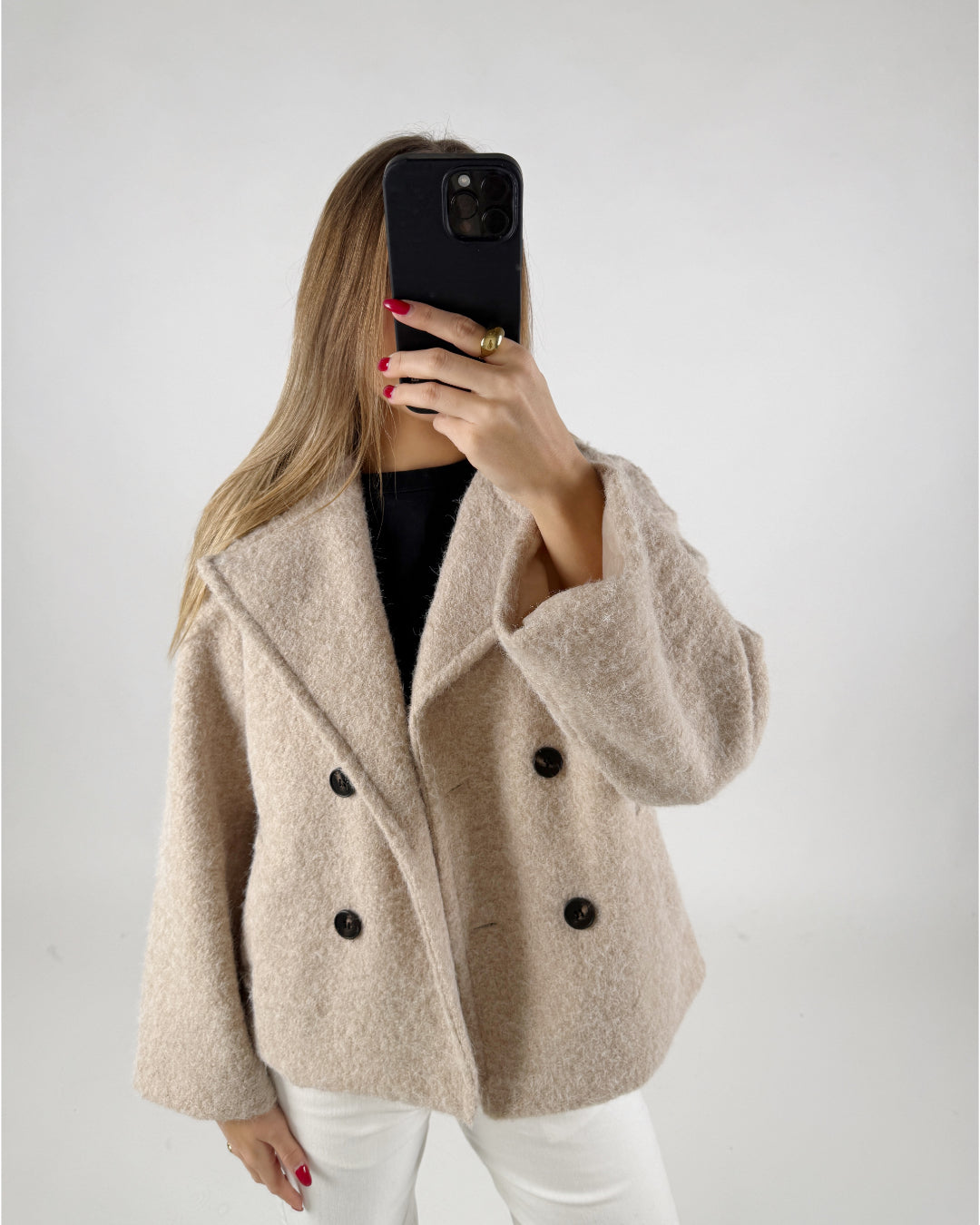 En person iført en beige frakke tager et billede med en sort telefon. Personen står i et neutralt, lysegråt rum.
