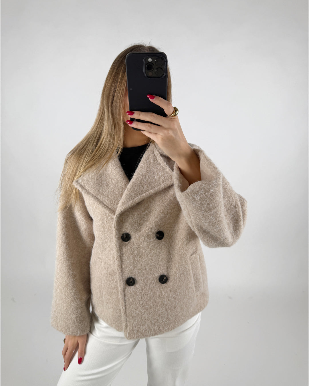 En person iført en beige frakke med sorte knapper holder en mobiltelefon op foran ansigtet. Omkring personen er der en simpel, lys baggrund.
