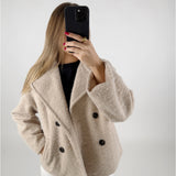 En person tager et selfie med en sort telefon. Personen bærer en beige frakke med sorte knapper og har rødt neglelak. Baggrunden er neutral med en hvid væg.