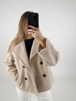 En person tager et selfie med en sort telefon. Personen bærer en beige frakke med sorte knapper og har rødt neglelak. Baggrunden er neutral med en hvid væg.