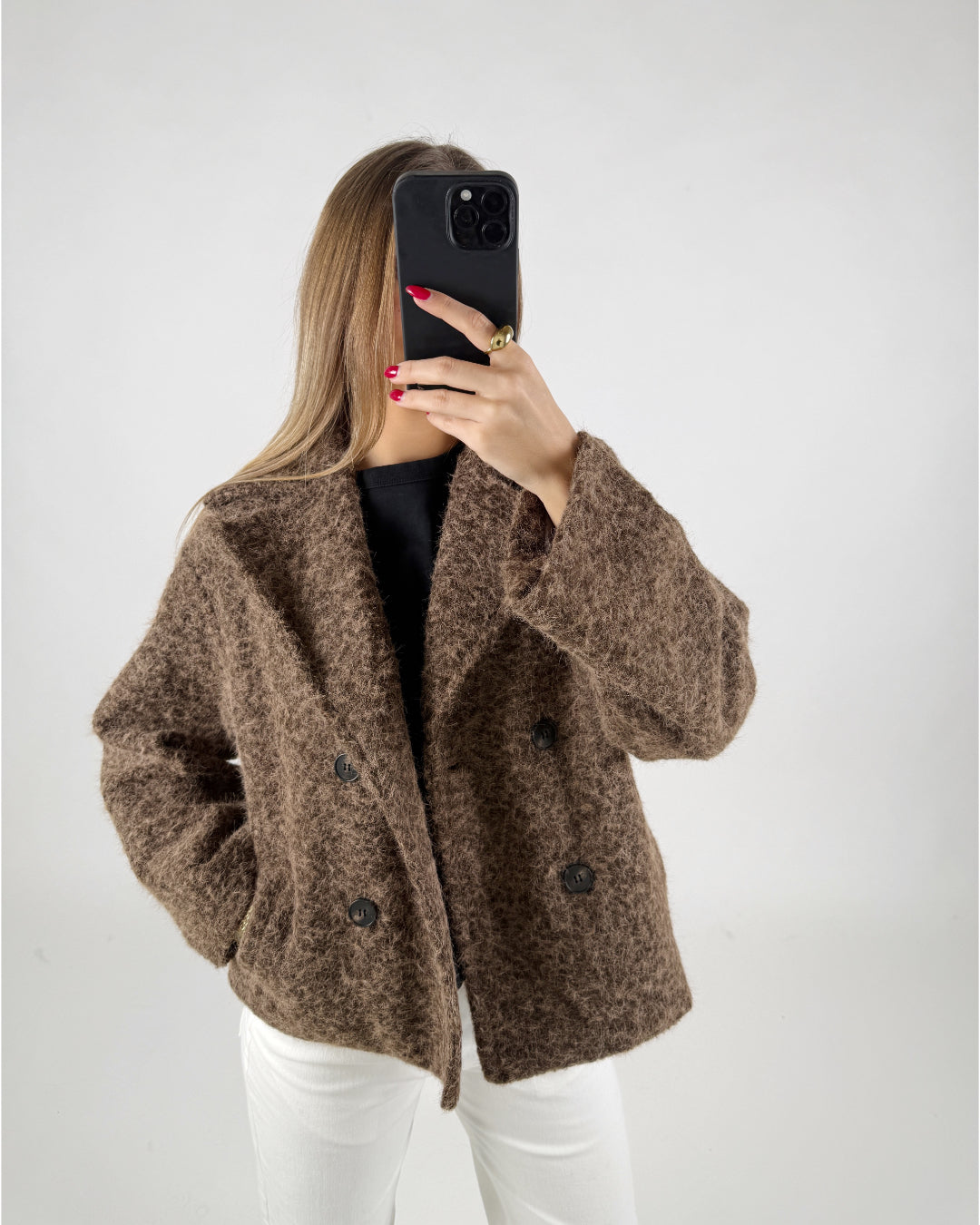 Kvinde iført brun, lodden jakke tager et selfie med sort telefon. Hun står i et lyst rum og har en sort trøje og hvide bukser på.