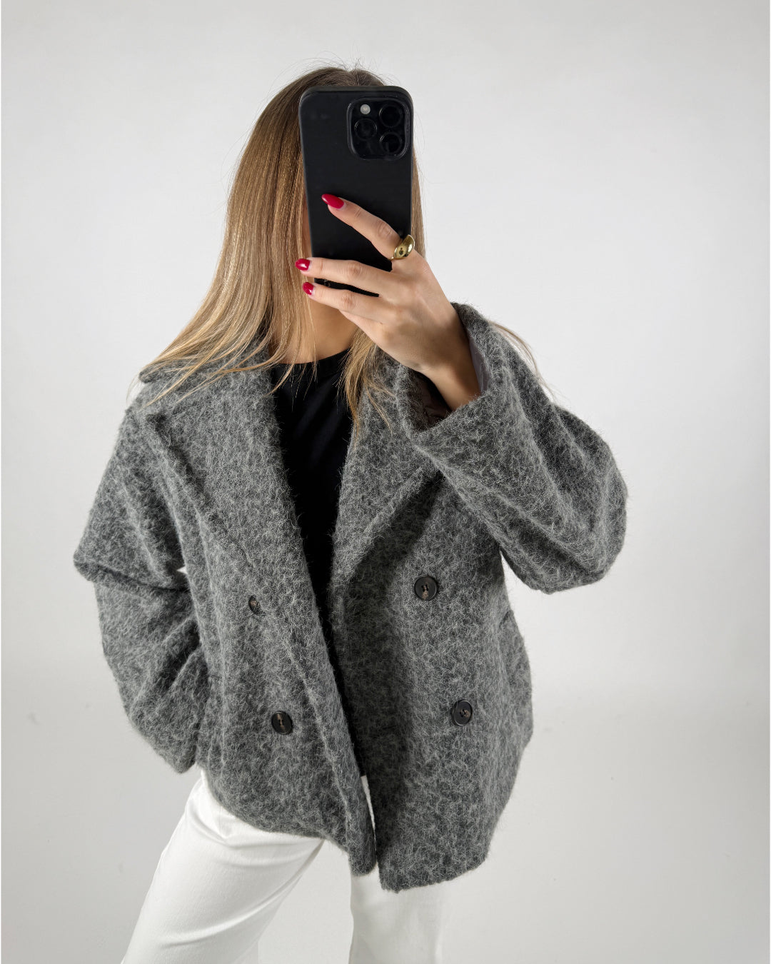 En person i grå frakke tager en selfie med en sort telefon. Personen har langt lyst hår, røde negle og står i et lyst rum.