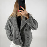 En person i grå frakke tager en selfie med en sort telefon. Personen har langt lyst hår, røde negle og står i et lyst rum.