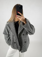 En person i grå frakke tager en selfie med en sort telefon. Personen har langt lyst hår, røde negle og står i et lyst rum.