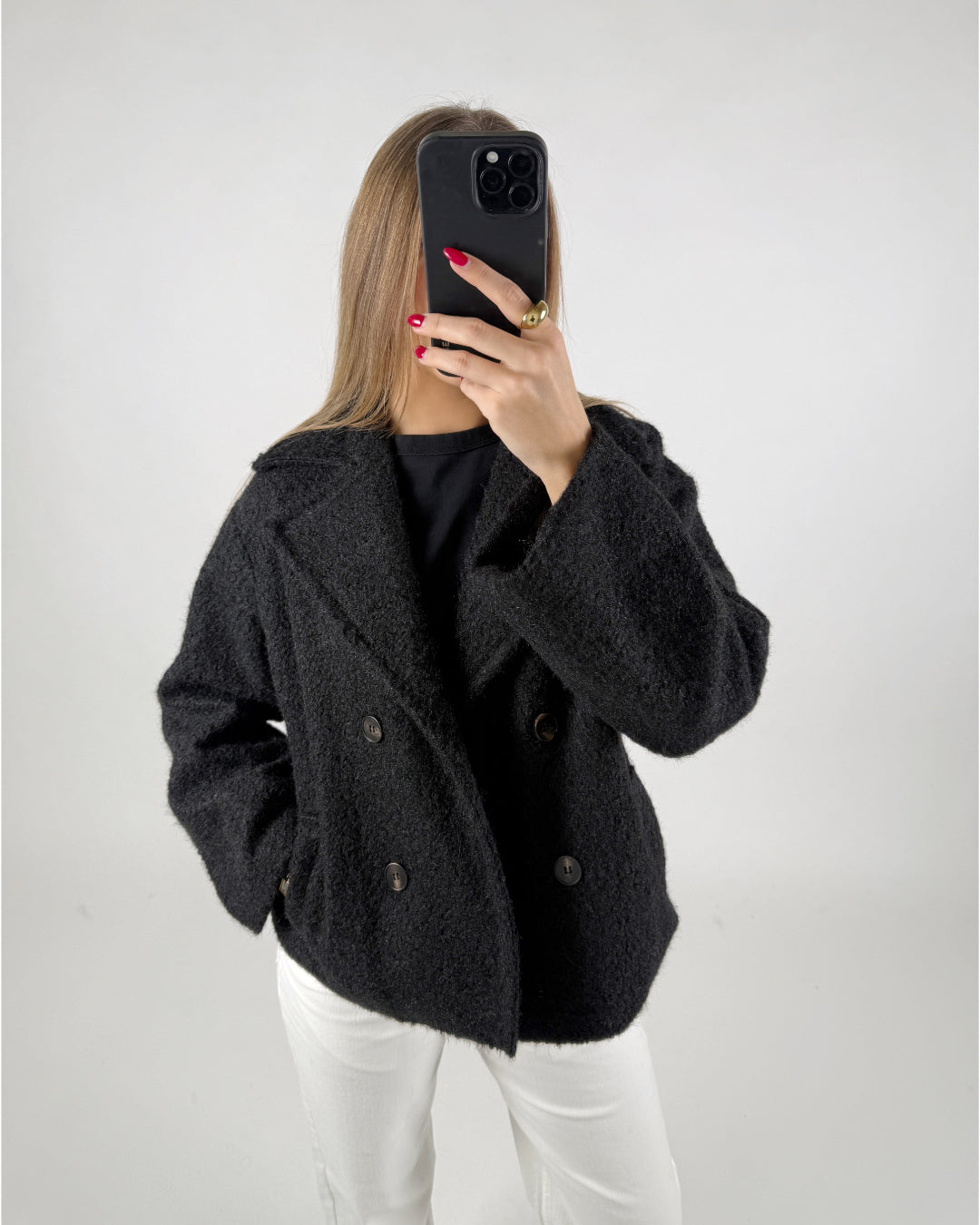 Person iført sort frakke tager et selfie med en sort smartphone i et lyst rum. Deres negle er røde, og de har en guldring på fingeren.