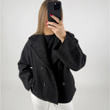 Person iført sort frakke tager et selfie med en sort smartphone i et lyst rum. Deres negle er røde, og de har en guldring på fingeren.