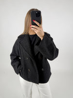 Person iført sort frakke tager et selfie med en sort smartphone i et lyst rum. Deres negle er røde, og de har en guldring på fingeren.