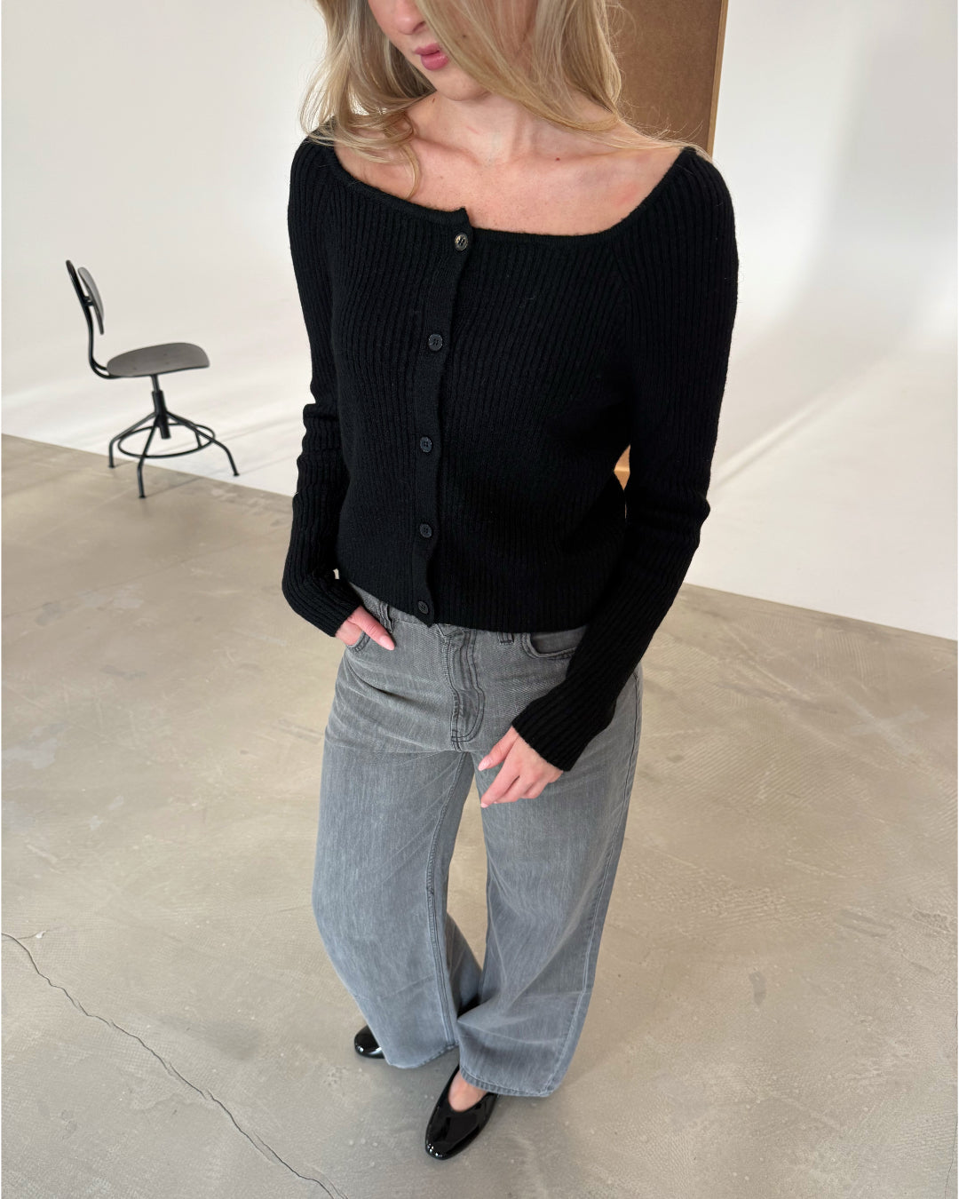 En person iført sort trøje og grå jeans står med hænderne i lommerne på et betongulv. I baggrunden ses en sort stol.