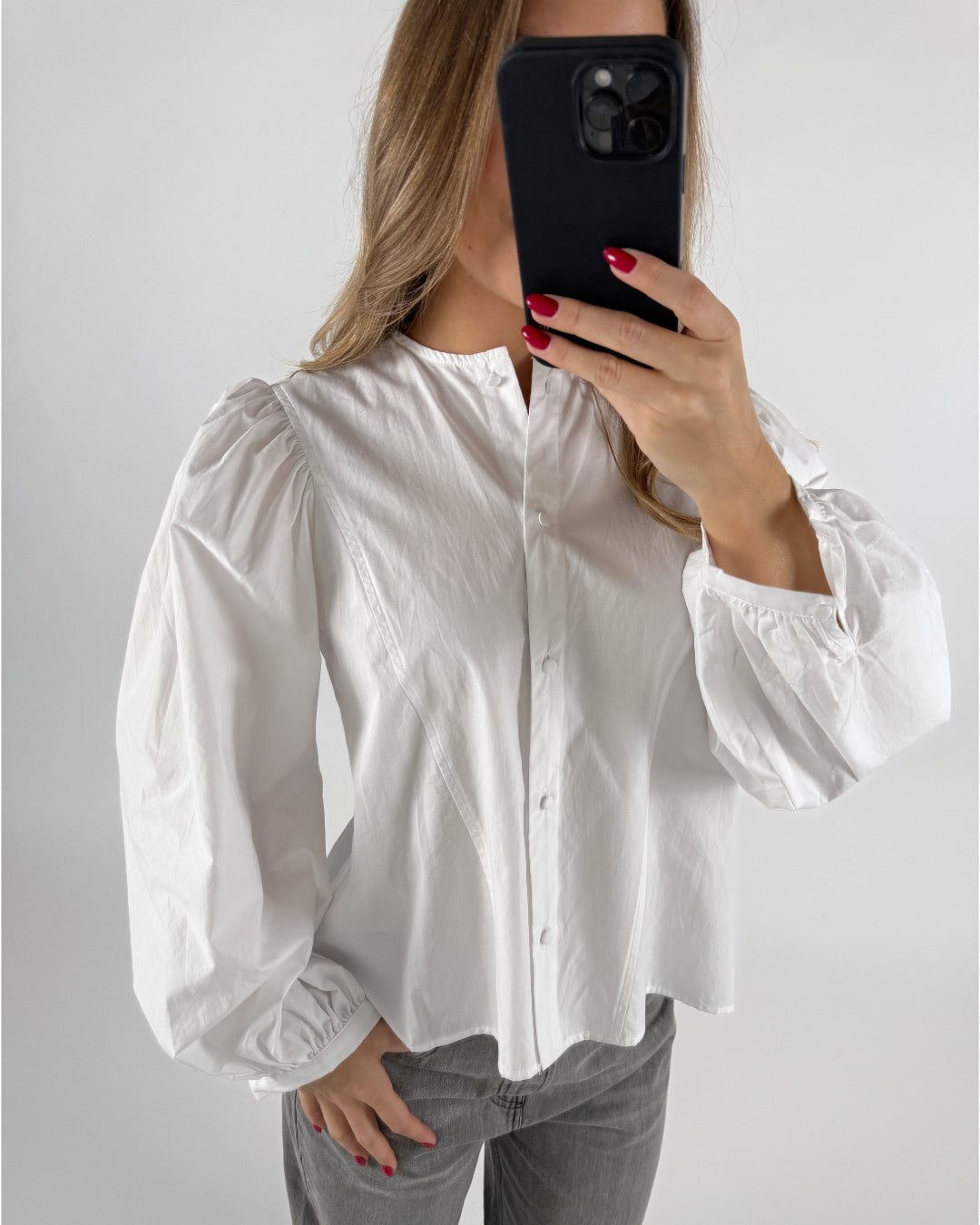 En person iført en hvid skjorte med pufærmer holder en sort smartphone foran ansigtet, omgivet af en neutral, hvid baggrund.